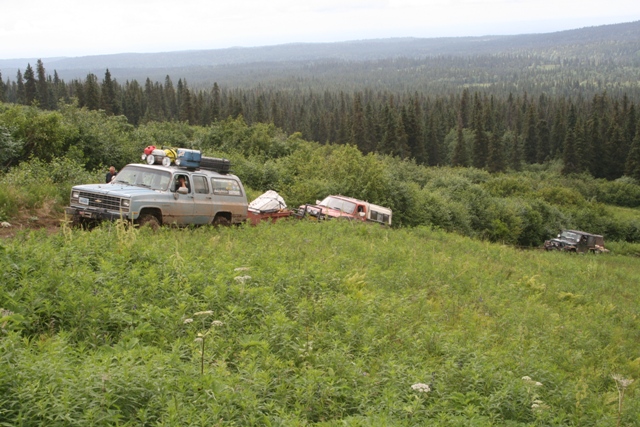 Light blue Chevrolet Suburban towing a trailer with a red vehicle, driving through a grassy, wooded area.