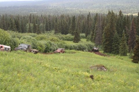 1990 Chevrolet Suburban - A convoy of off-road vehicles navigates a muddy trail through a dense forest and grassy hillside.