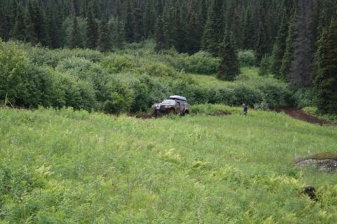 1990 Chevrolet Suburban - A lifted, camouflage-painted SUV with a roof rack is navigating through mud on a grassy slope. A person in a blue shirt stands nearby, obse…