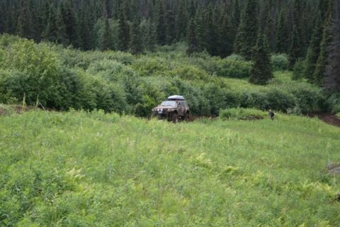 1990 Chevrolet Suburban - A lifted, mud-covered SUV with a roof rack navigates a grassy, wooded trail. A person stands in the background near the trees.