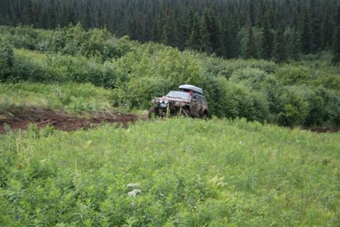 1990 Chevrolet Suburban - A muddy, dark-colored SUV with a roof-top carrier is navigating a grassy, uneven terrain with a dense forest in the background.