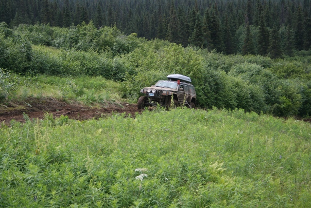 Muddy Jeep Cherokee with roof-top carrier on a dirt track in a wooded area.