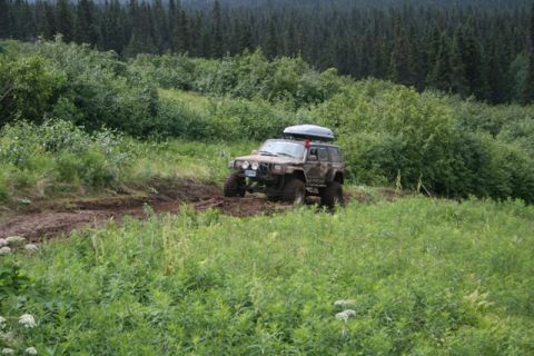 1990 Chevrolet Suburban - A muddy, lifted Jeep Cherokee is navigating a dirt track through a lush green forest. The vehicle is equipped with a roof-top cargo carrier…