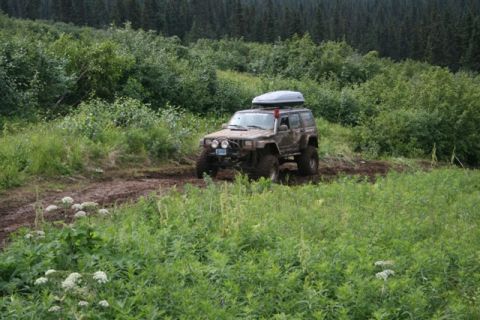 1990 Chevrolet Suburban - A muddy Jeep Cherokee XJ with a roof-top cargo box navigates a wet, rutted dirt trail through a lush green forest.