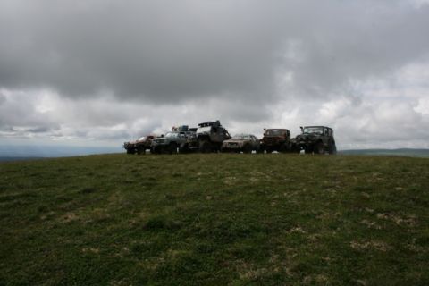 1990 Chevrolet Suburban - A line of six off-road vehicles are parked on a grassy hilltop under a cloudy sky. The vehicles are a mix of SUVs and modified trucks, with…