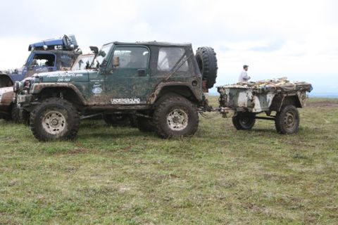 1990 Chevrolet Suburban - A muddy green Jeep Wrangler is parked on a grassy field, towing a small, camouflaged trailer. Another blue off-road vehicle is visible behi…