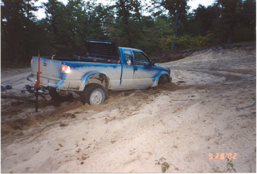 Blue and white extended-cab pickup truck stuck in deep sand, rear-side view, recovery jack nearby