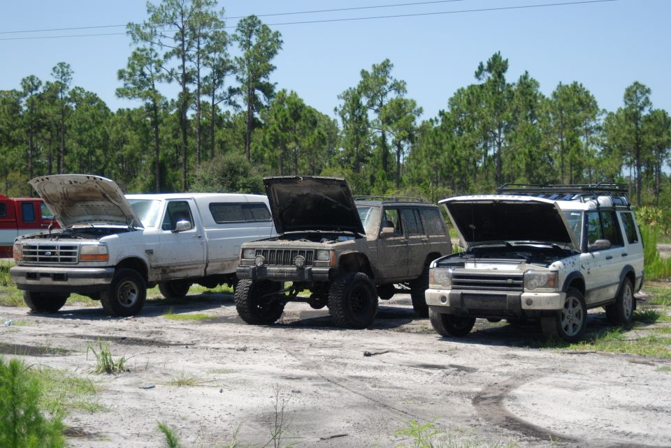 White Ford pickup, muddy lifted Jeep Cherokee, and Land Rover Discovery with hoods up on sandy lot