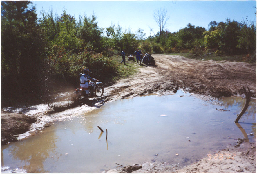 Dirt bike rider crossing a big mud puddle on an off-road trail, ATVs in background