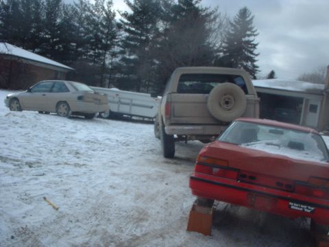 1986 Ford Bronco - A tan Ford Bronco from the 1980s is seen from the rear, parked in a snowy lot next to other vehicles.
