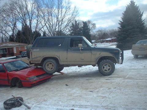 1986 Ford Bronco - A Ford Bronco is precariously positioned on top of a red car in a winter scene.