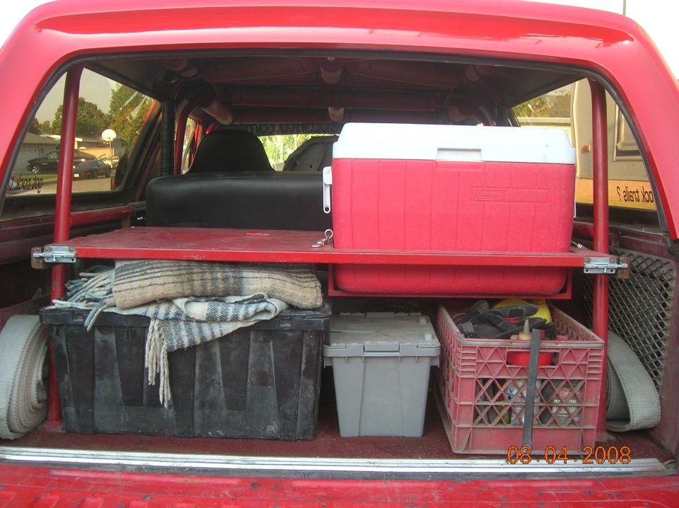 Red SUV cargo area with roll cage, red shelf holding a Coleman cooler, totes and gear