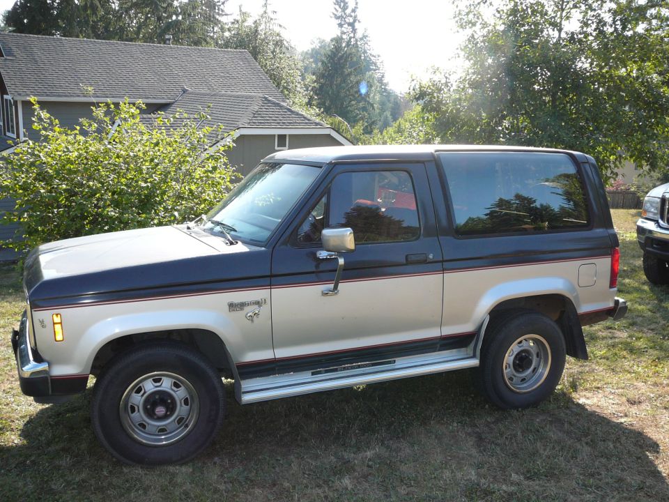 Two-tone blue and tan Ford Bronco II SUV, driver side view, parked on grass in front of a house