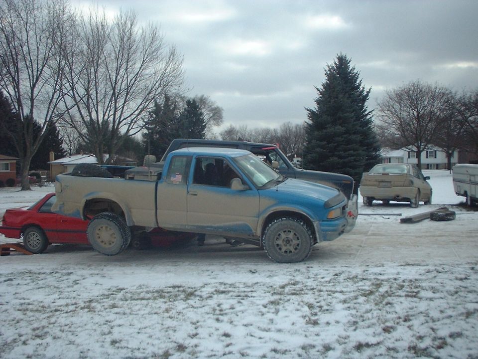 Blue and tan pickup truck parked next to a dark pickup truck, with a red car partially underneath the tan truck. Another tan car is in the background