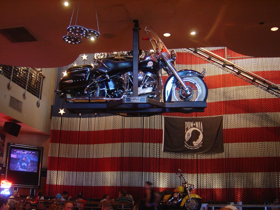 Black 1957 FLH Panhead motorcycle suspended, yellow motorcycle below, American flag backdrop.