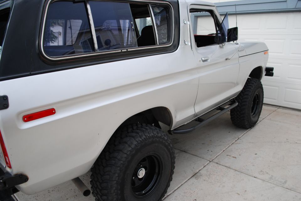 White 1978 Ford Bronco with black top, lifted on black wheels and mud tires, parked by garage doors