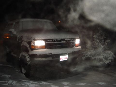 1996 Ford Bronco - A Ford truck is covered in snow, with its headlights on, emitting exhaust in the cold night air.