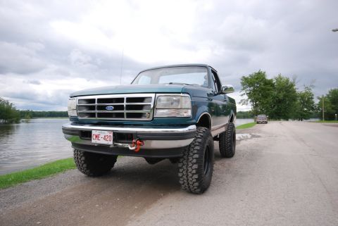 1996 Ford Bronco - A lifted green Ford F-Series truck sits by the water under a cloudy sky.