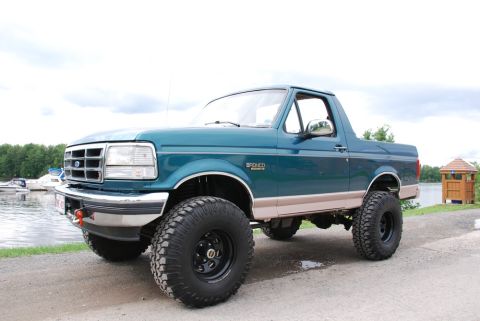 1996 Ford Bronco - A teal 1996 Ford Bronco with beige trim and large off-road tires sits parked near a body of water under a cloudy sky.