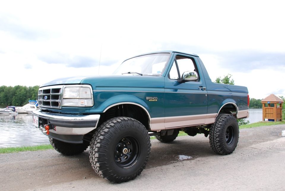 Teal 1996 Ford Bronco with beige trim and large off-road tires parked by a lake