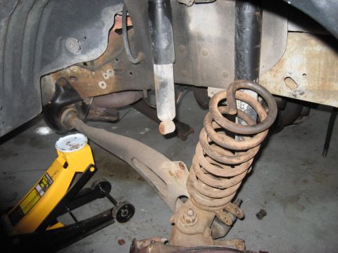 1991 Ford Bronco - Vehicle suspension components are visible, including a shock absorber and a rusty coil spring, with a jack supporting the frame.