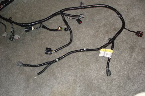 2007 Ford Mustang - A black wiring harness lies on a gray carpeted surface, a yellow identification tag and various electrical connectors.