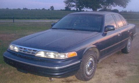 1997 Ford Crown Victoria - A dark blue sedan sits in a grassy field as the sun sets, reflecting on its windows.