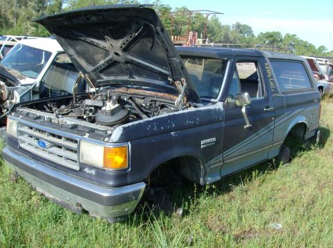 1989 Ford Bronco - A dark blue Ford Bronco sits in a junkyard with its hood open and missing front tires.