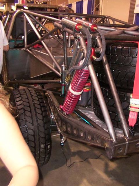 1984 Ford Bronco - Rear corner of a bare tube-chassis desert race truck on display at an indoor show, showing a cantilever suspension with a red coil spring o…