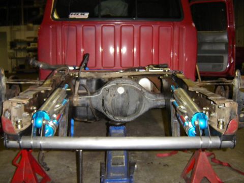 1984 Ford Bronco - Rear view of a red Ford truck on jack stands with the bed removed, exposing a bare rear axle housing supported on a blue floor jack. A cust…
