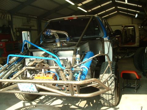 1984 Ford Bronco - Rear 3/4 view of a black prerunner truck under construction in a metal shop, with the bed replaced by a bare tube-chassis rear section carr…
