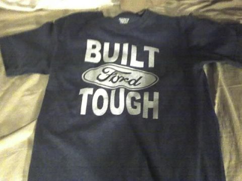 1996 Ford Bronco - A dark navy short-sleeve t-shirt laid flat on a bed, printed with the silver 'BUILT FORD TOUGH' logo including the oval Ford emblem.