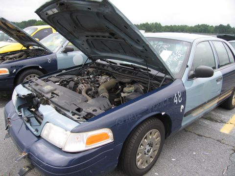 2001 Ford Crown Victoria - A two-tone Ford Crown Victoria with its hood open, revealing the engine compartment, sits in a lot with other vehicles.