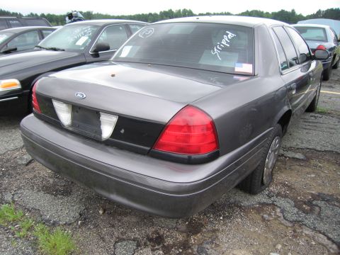 2001 Ford Crown Victoria - A gray 2001 Ford Crown Victoria is parked in a lot with other vehicles, showing a flat tire and 'stripped' written on the rear window.
