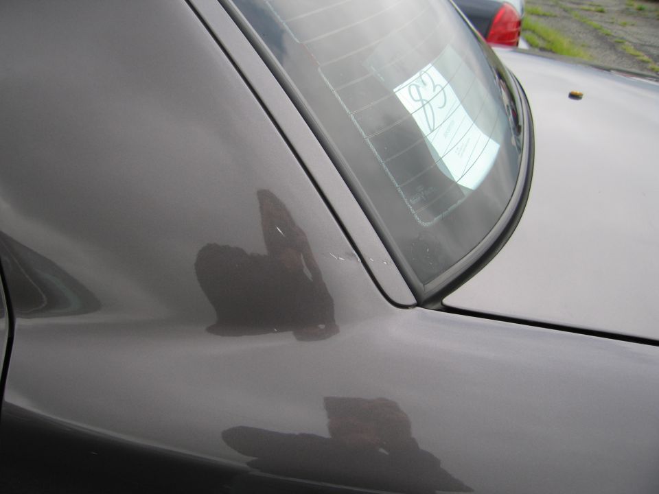 Reflection of person in rear quarter panel and rear window of dark sedan. Paper with '83' visible on rear window