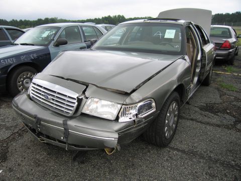 2001 Ford Crown Victoria - A damaged Ford Crown Victoria sits in a lot with its hood and doors open, next to a State Police vehicle.