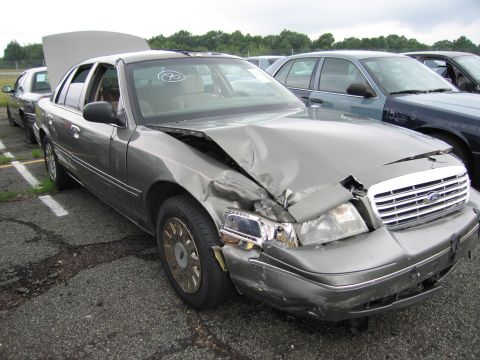 2001 Ford Crown Victoria - A damaged 2001 Ford Crown Victoria is parked in a lot with its hood open, showing significant front-end collision damage.