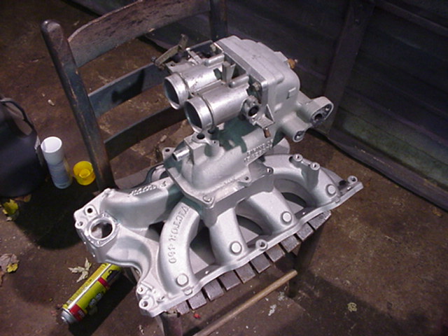 Silver-painted V-8 aluminum intake manifold with throttle body on a wooden chair