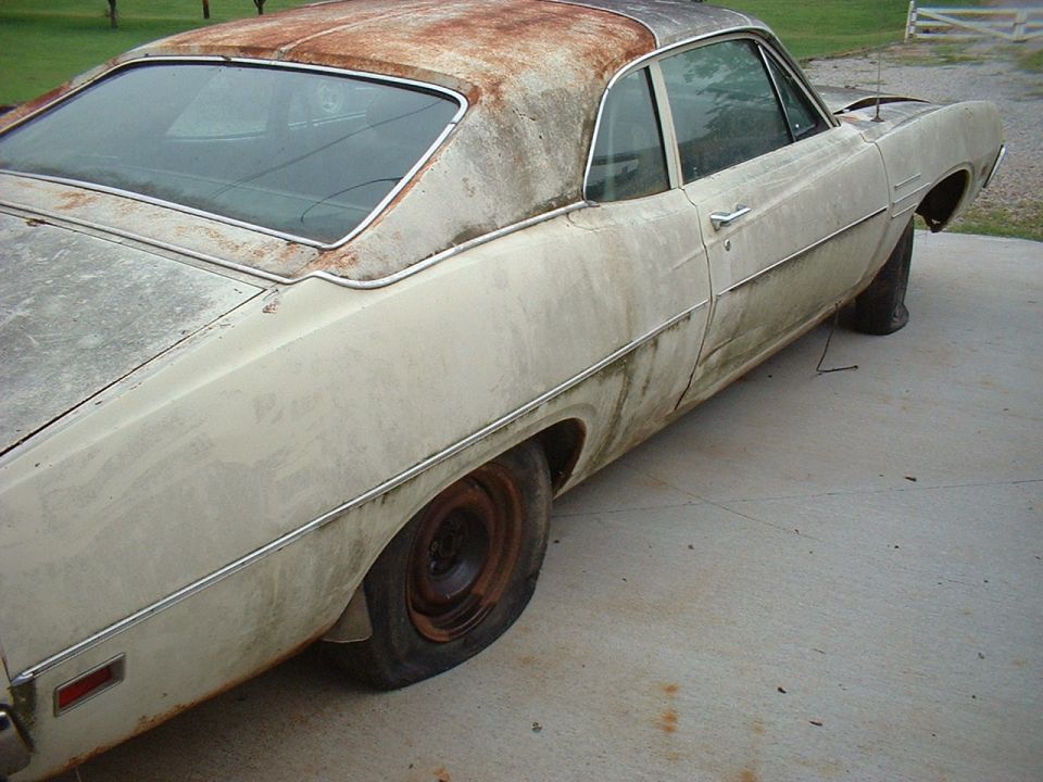Weathered cream two-door project car with rusty roof and flat tire on bare steel wheel, parked on concrete