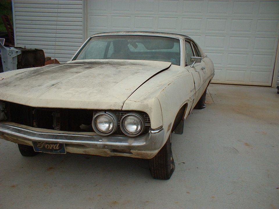 Dusty white Ford fastback project car with missing grille center, front resting on wood stumps