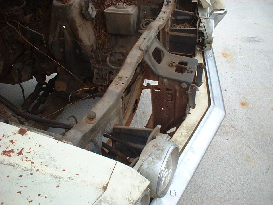 Stripped front end of old white car showing rusty core support and round headlight bucket