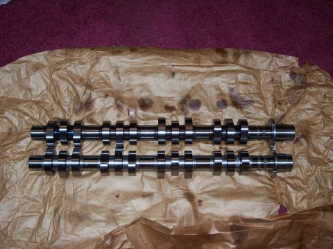 2007 Ford Mustang - Two camshafts are laid out on brown paper, with a dark red carpet visible in the background. The camshafts are made of polished metal and h…