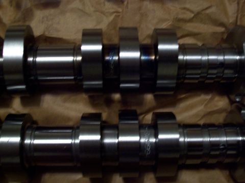 2007 Ford Mustang - Two shiny metal camshafts are laid out on brown paper. Each camshaft has multiple lobes and appears to be a performance part.