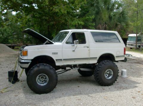 1989 Ford Bronco - A lifted, white 1980s Ford Bronco with large off-road tires is parked on a gravel surface with the hood open. A black battery charger is co…