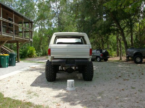 1989 Ford Bronco - The rear view of a lifted, white Ford Bronco is shown parked on a gravel driveway next to a wooden deck. Another SUV and a pickup truck are…