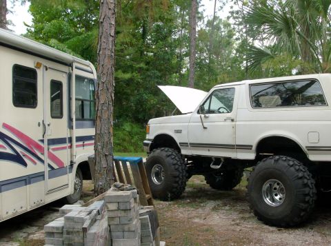1989 Ford Bronco - A white, lifted Ford Bronco with oversized tires is parked next to an RV in a wooded area. The hood of the Bronco is open, and a stack of b…