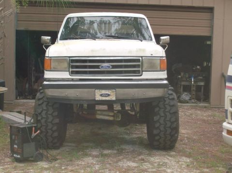 1989 Ford Bronco - Front view of a lifted white Ford Bronco with large off-road tires parked in front of an open garage door. A black solar battery charger is…