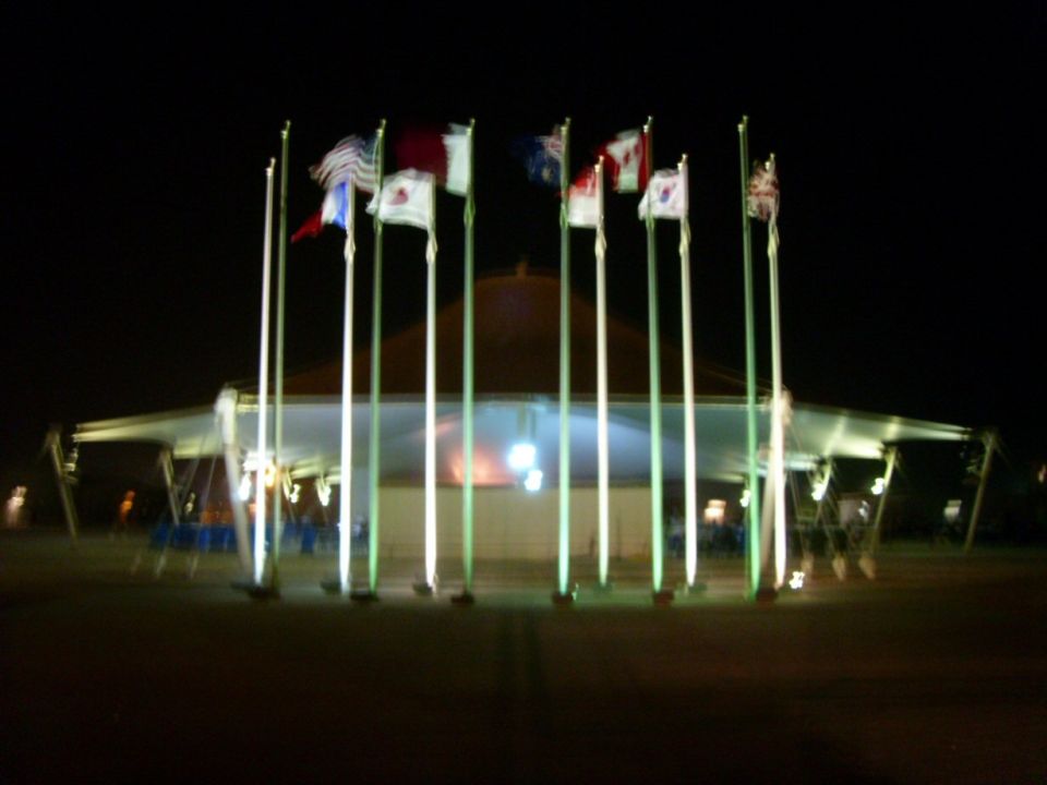 Row of illuminated international flags on poles in front of a lit tent at night