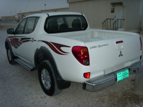 1978 Ford Bronco - Rear three-quarter view of a white Mitsubishi L200 Sportero double-cab pickup on a gravel lot, with red tribal flame graphics along the bed…