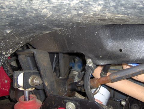 1978 Ford Bronco - The underside of a vehicle shows a black plastic fender liner above rusty metal components and a black rubber hose secured with a clamp.